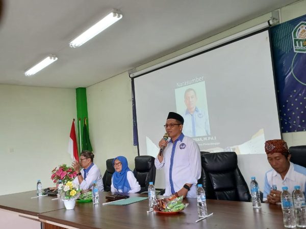 Empat orang duduk di meja konferensi, satu pria berdiri berbicara dengan mikrofon. Meja berisi air mineral, camilan, dan bunga. Layar proyektor di belakang menampilkan foto dan tulisan “Narasumber”. Ada bendera Indonesia dan dua bendera hijau di sudut ruangan. Suasana formal, seperti seminar.