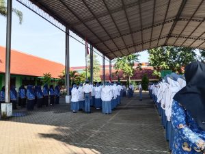 Suasana upacara bendera yang berlangsung di GOR  MAN 2 Probolinggo. Barisan siswa berseragam putih-abu-abu berdiri tegak menghadap ke depan, di sebelah kiri terdapat barisan guru wanita berseragam batik biru, dan di sebelah kanan sebagian guru wanita juga terlihat. Bendera Merah Putih telah dikibarkan di tiang di tengah lapangan. Latar belakang menunjukkan gedung sekolah beratap merah dan pepohonan di bawah langit cerah.